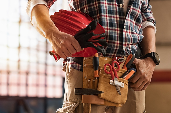 Construction worker with a tool belt holding a red hard hat and gloves.