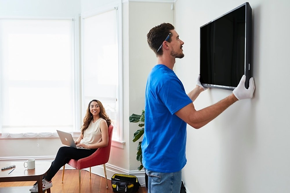 Technician mounting a TV on the wall while a woman watches from a chair.