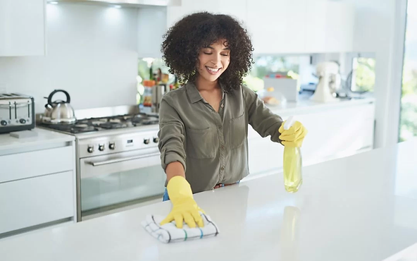 Smiling woman cleaning a kitchen countertop with a spray bottle and cloth.